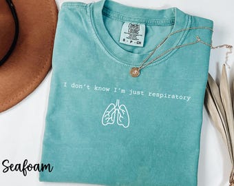 Pode incluir: Camiseta verde-água com o texto "I don't know I'm just respiratory" e um gráfico branco de pulmões. Um colar de ouro com um pingente está sobre a camiseta. A camiseta é da marca Comfort Colors.