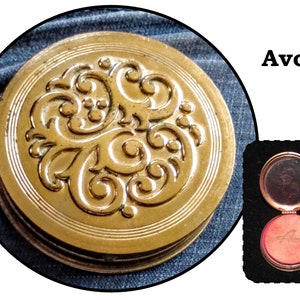 May include: A gold compact with an ornate floral design. The compact is open, revealing a red, velvet-lined interior with the word "Avon" embossed in gold.