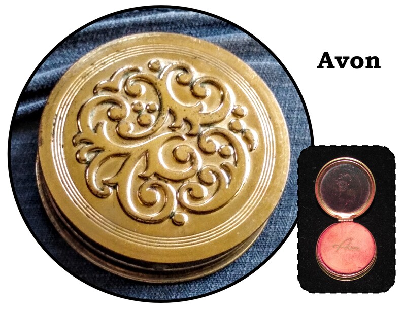 May include: A gold compact with an ornate floral design. The compact is open, revealing a red, velvet-lined interior with the word "Avon" embossed in gold.
