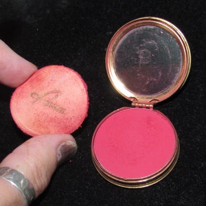 May include: A vintage compact with a gold-colored metal case and a pink blush. The compact has a mirror and a  velvet-lined lid with the word "Avon" printed in gold.