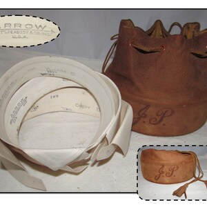 May include: A brown leather drawstring bag with a tassel and the initials "J.P." embroidered on the front. The bag is sitting on a white surface next to a white fabric collar with the brand name "Arrow" printed on it.