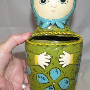 Pu&ograve; includere: Un vaso da fiori in ceramica a forma di bambola con un corpo verde, accenti blu e gialli e un design floreale blu e bianco. La bambola ha un foulard blu e capelli gialli.