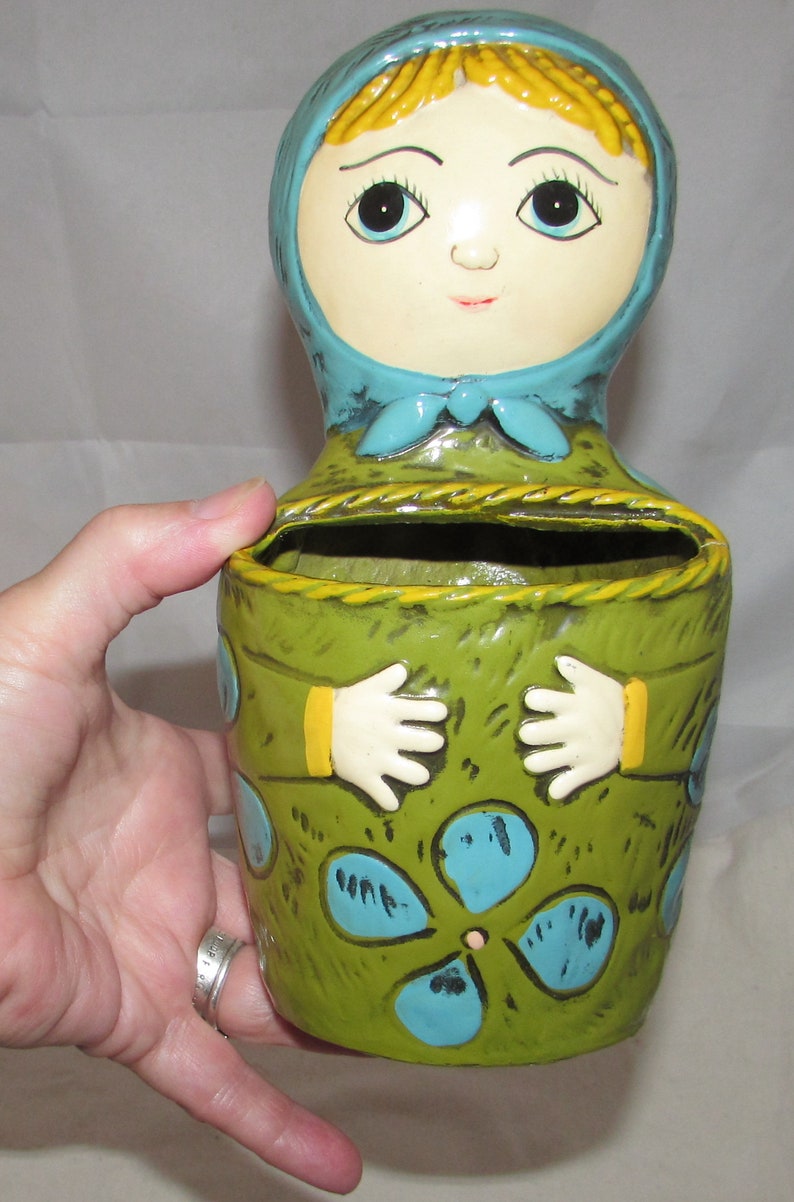 Pu&ograve; includere: Un vaso da fiori in ceramica a forma di bambola con un corpo verde, accenti blu e gialli e un design floreale blu e bianco. La bambola ha un foulard blu e capelli gialli.