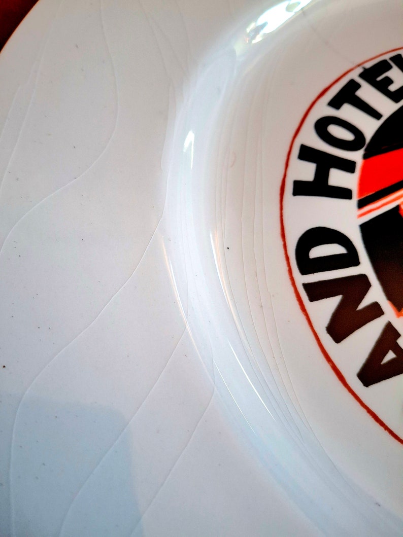 Op de afbeelding: Witte keramische plaat met een zwart en rood cirkelontwerp met de tekst "Grand Hotel" in zwarte letters.