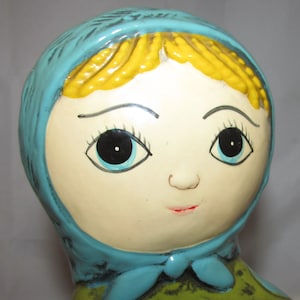 Pu&ograve; includere: Una bambola di ceramica con un foulard blu, capelli gialli e tratti dipinti. La bambola ha grandi occhi neri con iride blu e un sorriso dipinto.