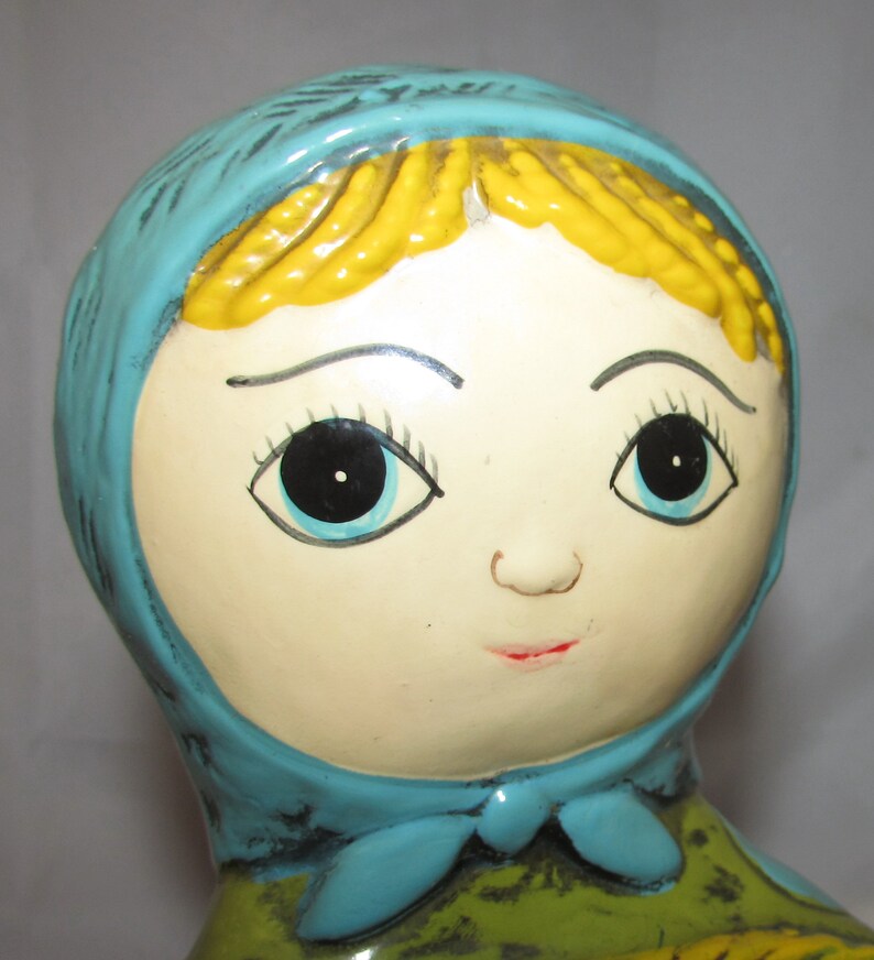 Pu&ograve; includere: Una bambola di ceramica con un foulard blu, capelli gialli e tratti dipinti. La bambola ha grandi occhi neri con iride blu e un sorriso dipinto.