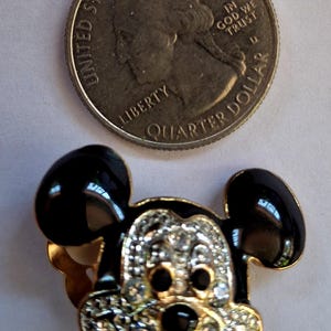 Puede incluir: Broche de Mickey Mouse en tono dorado con orejas de esmalte negro y una cara incrustada con pedrer&iacute;a transparente. Una moneda de cuarto de d&oacute;lar estadounidense est&aacute; al lado del broche para comparar el tama&ntilde;o. El broche tiene una boca roja.