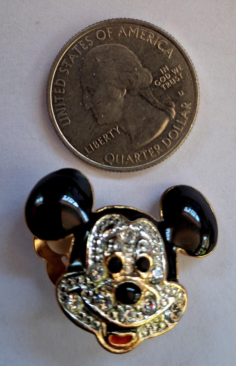 Puede incluir: Broche de Mickey Mouse en tono dorado con orejas de esmalte negro y una cara incrustada con pedrer&iacute;a transparente. Una moneda de cuarto de d&oacute;lar estadounidense est&aacute; al lado del broche para comparar el tama&ntilde;o. El broche tiene una boca roja.