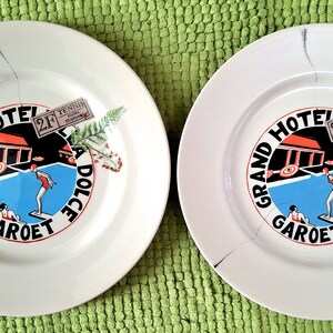 Op de afbeelding: Twee witte keramische borden met een blauw, rood en zwart ontwerp met de tekst "Grand Hotel Garot Dolce Vita" en een klein kaartje met de tekst "Tennis 2F".