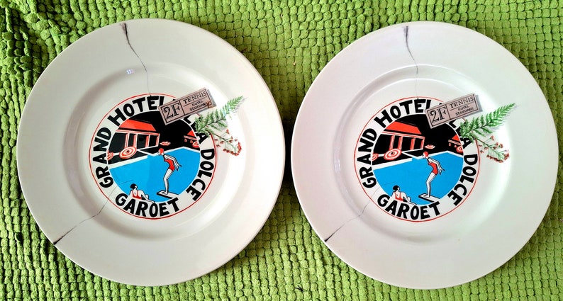 Op de afbeelding: Twee witte keramische borden met een blauw, rood en zwart ontwerp met de tekst "Grand Hotel Garot Dolce Vita" en een klein kaartje met de tekst "Tennis 2F".