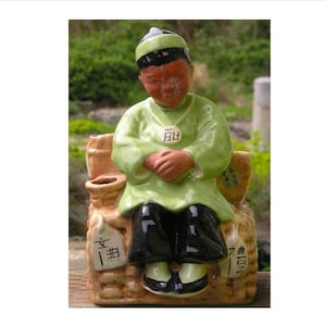 May include: A ceramic figurine of a man wearing a green robe and black pants sitting on a woven basket. The figurine has a brown face and black hair. The basket has Japanese characters on it.
