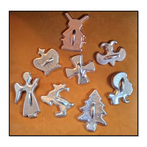 May include: A collection of vintage metal cookie cutters in various shapes, including a windmill, crown, cross, angel, reindeer, Christmas tree, and a boat. The cutters are silver-colored and arranged on a brown surface.