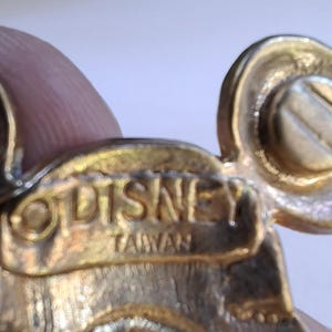Puede incluir: Primer plano de un pin dorado de la marca Disney. El pin presenta un dise&ntilde;o de cabeza de Mickey Mouse con un detalle de tornillo. La palabra "DISNEY" y "TAIWAN" est&aacute;n grabadas en el pin.