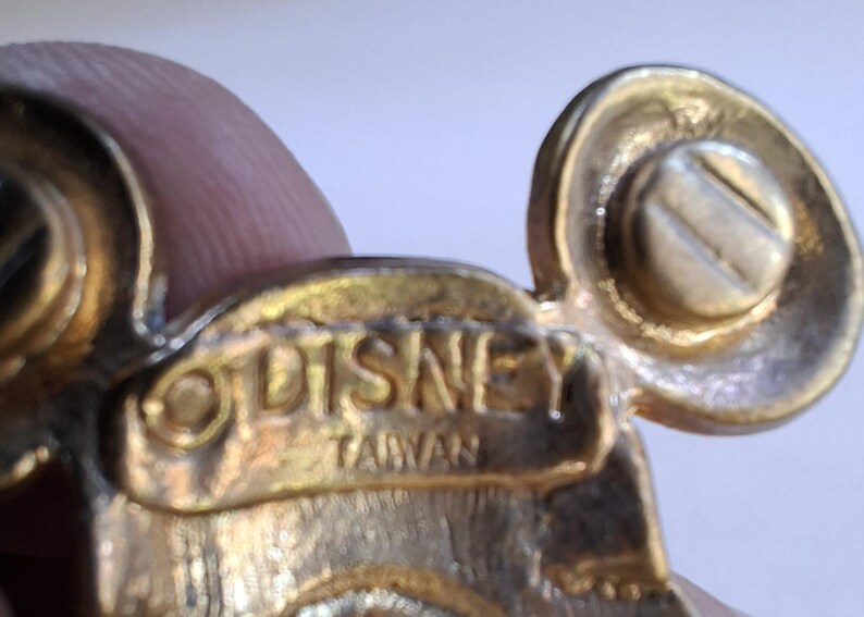 Puede incluir: Primer plano de un pin dorado de la marca Disney. El pin presenta un dise&ntilde;o de cabeza de Mickey Mouse con un detalle de tornillo. La palabra "DISNEY" y "TAIWAN" est&aacute;n grabadas en el pin.