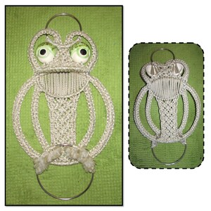 May include: A white macrame frog wall hanging with green eyes and a silver ring. The frog is hanging on a green background.