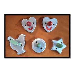 May include: A collection of vintage cookie cutters in various shapes, including hearts, a star, a chicken, and a circle. Each cutter is made of silver metal and features a colorful knob and painted details. The cutters are arranged on a brown surface.