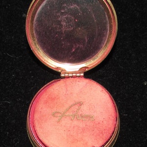 May include: A gold compact with a round mirror and a red velvet interior. The interior has the word "Avon" written in gold script.