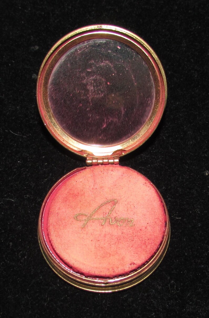 May include: A gold compact with a round mirror and a red velvet interior. The interior has the word "Avon" written in gold script.