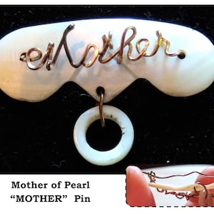 May include: A white mother of pearl pin with a gold wire inscription that reads "Mother". The pin has a white mother of pearl ring attached to the bottom.
