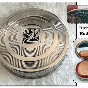 May include: Silver compact with a black and white design on the lid. The compact is open and shows a red velvet cushion with a mirror on the top. The compact is labeled "MARVELOUS RICHARD HUDNUT MADE IN U.S.A."