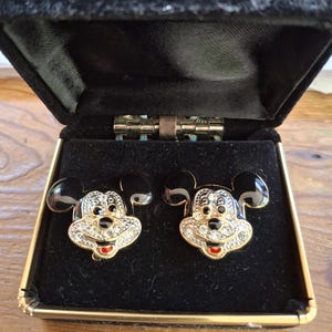 Puede incluir: Un par de gemelos de Mickey Mouse en una caja de terciopelo negro. Los gemelos presentan una cara dorada con pedrer&iacute;a transparente, orejas negras y boca roja. La caja tiene un ribete dorado.