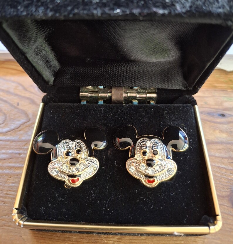 Puede incluir: Un par de gemelos de Mickey Mouse en una caja de terciopelo negro. Los gemelos presentan una cara dorada con pedrer&iacute;a transparente, orejas negras y boca roja. La caja tiene un ribete dorado.