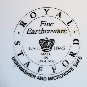 Op de afbeelding: Een zwart-witte ronde stempel met de tekst "ROYAL STAFFORD Fine Earthenware EST. 1845 MADE IN ENGLAND" en een kroonafbeelding. De tekst "DISHWASHER AND MICROWAVE SAFE" staat rond de buitenkant van de cirkel.