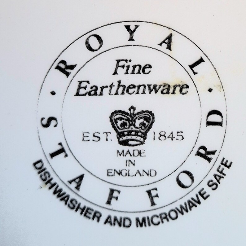 Op de afbeelding: Een zwart-witte ronde stempel met de tekst "ROYAL STAFFORD Fine Earthenware EST. 1845 MADE IN ENGLAND" en een kroonafbeelding. De tekst "DISHWASHER AND MICROWAVE SAFE" staat rond de buitenkant van de cirkel.