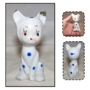 May include: A white porcelain figurine of a cat with blue polka dots and black eyelashes. The cat is sitting with its head tilted slightly to the side.