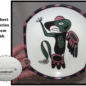 May include: A white ceramic dish with a black and pink border. The dish features a hand-painted green, black, and pink totem pole design. The bottom of the dish is marked with "Lambert Potteries #45. SKOOKUM. G."