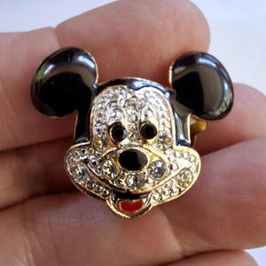 Puede incluir: Broche de Mickey Mouse dorado con orejas negras y una cara incrustada con pedrer&iacute;a transparente. El broche presenta una nariz, ojos negros y una boca roja. Es un dise&ntilde;o cl&aacute;sico de personaje de Disney.