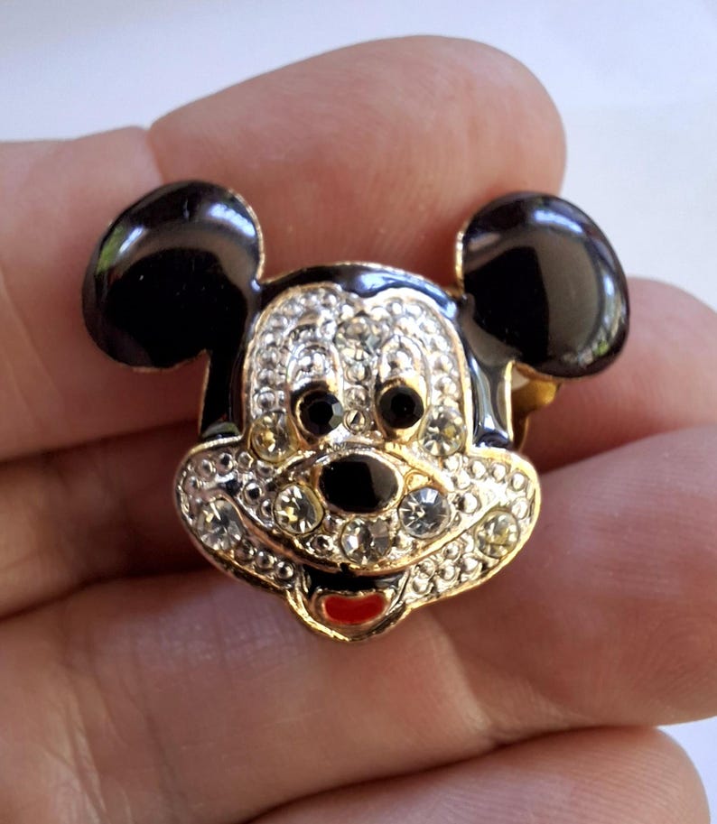 Puede incluir: Broche de Mickey Mouse dorado con orejas negras y una cara incrustada con pedrer&iacute;a transparente. El broche presenta una nariz, ojos negros y una boca roja. Es un dise&ntilde;o cl&aacute;sico de personaje de Disney.