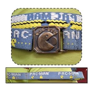 May include: A Pac-Man themed belt with a bronze-colored buckle featuring the Pac-Man character. The blue belt has the words "PAC-MAN" and yellow Pac-Man ghosts embroidered on it. The top of the belt has the words "EAT DOTS" in white and yellow.