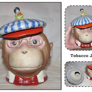 May include: A ceramic tobacco jar shaped like a monkey's head wearing a blue and white striped hat, red glasses, and a red bow tie. The lid features a small figurine of a child wearing a green dress.