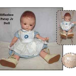 May include: A vintage Effanbee Patsy Jr. doll wearing a blue and white striped dress with a white apron and a blue bird design. The doll is sitting on a white surface and has brown hair and blue eyes. The doll's feet are wearing white shoes with brown soles. The doll's name is printed on the bottom of the doll's foot.