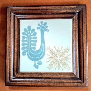 May include: A square ceramic tile set within a dark brown wooden frame. The tile features a light blue stylized bird graphic on the left side and a gold-colored geometric wheat-like pattern on the right side against a cream-colored background.