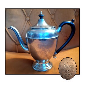 May include: A silver-colored teapot with a black handle and a black finial on the lid. The teapot has a curved spout and sits on a pedestal base. A small, circular emblem with the text "TP OR COPPER" is visible.