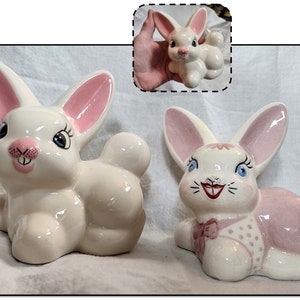 May include: Two ceramic bunny figurines. One is white with pink ears and a pink nose. The other is white with pink ears, a pink nose, and a pink dress with white polka dots.