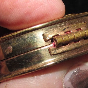 May include: A gold-toned compact with a hinged lid and a brass hinge. The compact is closed and held in a hand.