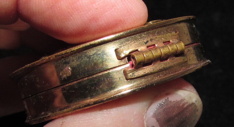 May include: A gold-toned compact with a hinged lid and a brass hinge. The compact is closed and held in a hand.