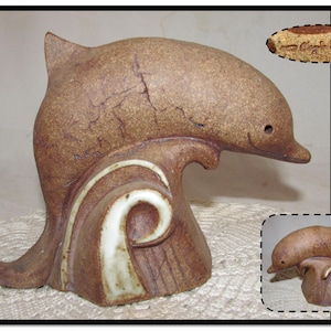 May include: A brown ceramic dolphin sculpture leaping out of the water. The dolphin is glazed in a brown and white color scheme. The sculpture is signed "Craft Fair" on the base.