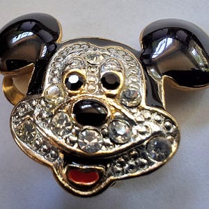 Puede incluir: Broche con forma de Mickey Mouse con orejas y contorno de la cara de esmalte negro. La cara est&aacute; adornada con pedrer&iacute;a transparente y negra, una boca roja y detalles met&aacute;licos dorados.