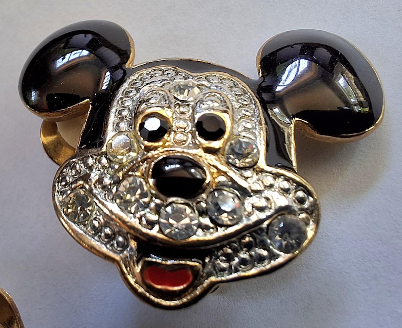 Puede incluir: Broche con forma de Mickey Mouse con orejas y contorno de la cara de esmalte negro. La cara est&aacute; adornada con pedrer&iacute;a transparente y negra, una boca roja y detalles met&aacute;licos dorados.