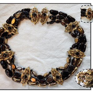 May include: A gold and black necklace with a large, ornate clasp. The necklace is made of oval-shaped beads and features a repeating pattern of gold accents. The clasp is marked with the word "Eglon".