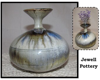 May include: A white ceramic vase with brown and blue glaze. The vase has a wide, round body and a narrow neck. The vase is sitting on a white lace tablecloth. The text "Jewell Pottery" is visible in the lower right corner of the image.
