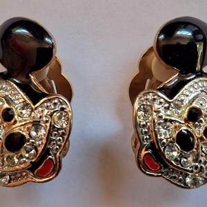 Puede incluir: Un par de pendientes de clip con forma de Mickey Mouse. Los pendientes presentan cabezas y orejas de esmalte negro, con la cara y el cuerpo delineados en dorado e incrustados con pedrer&iacute;a transparente. Se ven detalles en rojo.
