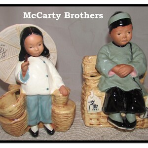 May include: Two vintage ceramic figurines of a boy and girl dressed in traditional Chinese clothing. The girl holds a white parasol and both figures are sitting on baskets.