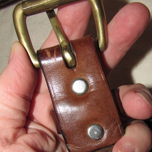 May include: A brown leather belt with a brass buckle. The buckle is closed and the belt is held in a hand.
