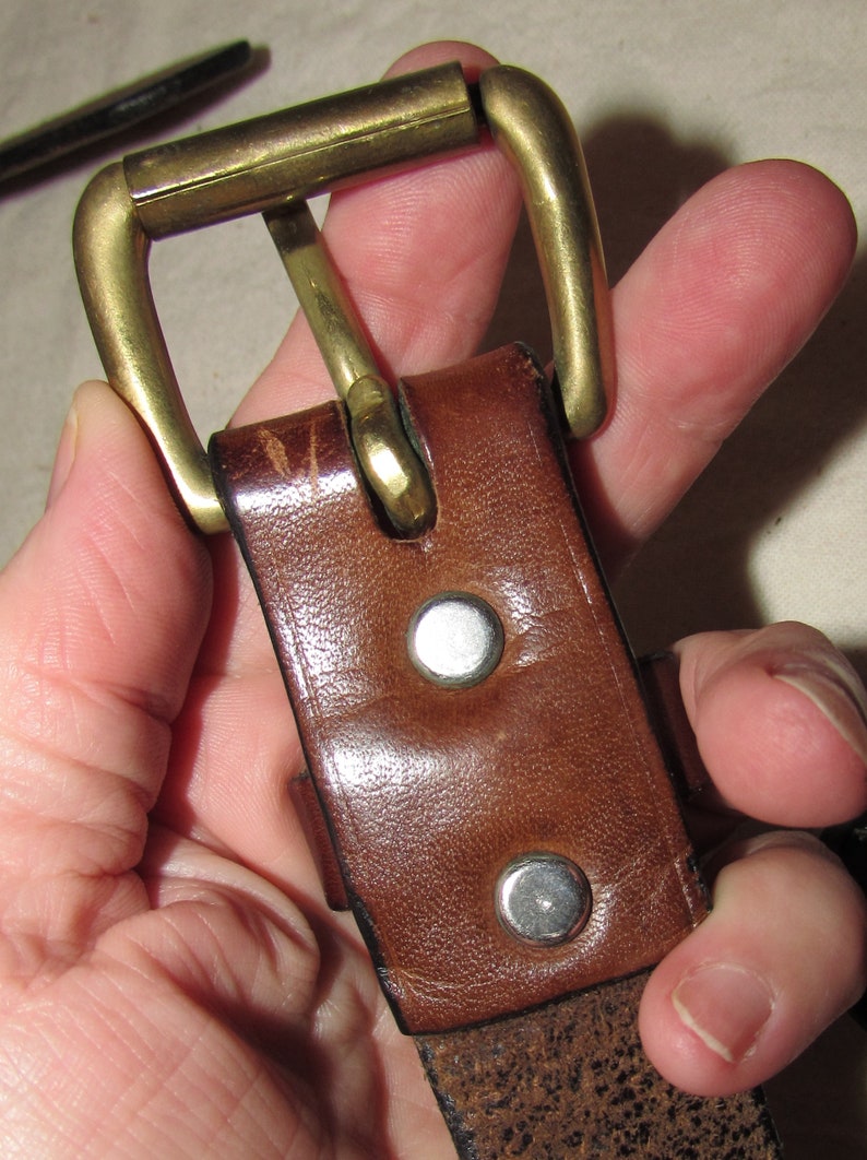 May include: A brown leather belt with a brass buckle. The buckle is closed and the belt is held in a hand.