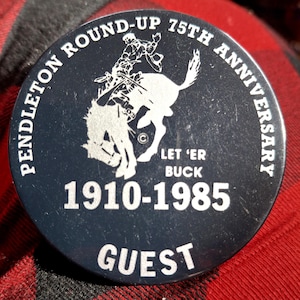 May include: Black and white button with a cowboy riding a bucking bronco. The button says "Pendleton Round-Up 75th Anniversary" and "Let 'er Buck" with the years "1910-1985" and the word "Guest".